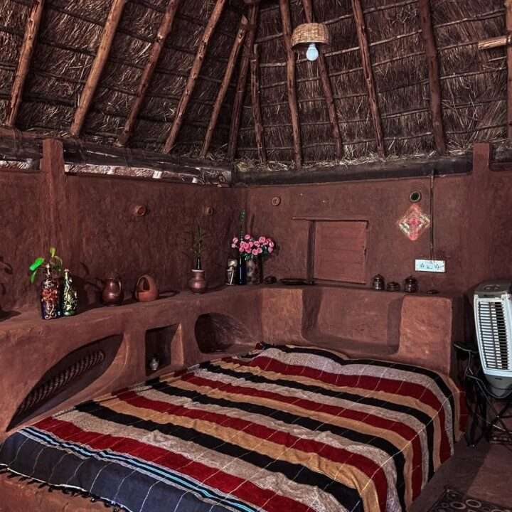 AuraKalari : The AuraKalari accommodation offers a rustic bedroom with a thatched roof and walls made of mud or clay. A double bed is covered with a multicolored striped blanket. Shelves built into the walls hold various clay pots and decorative items. A small window above the bed allows natural light, while an air cooler is placed on the right side.